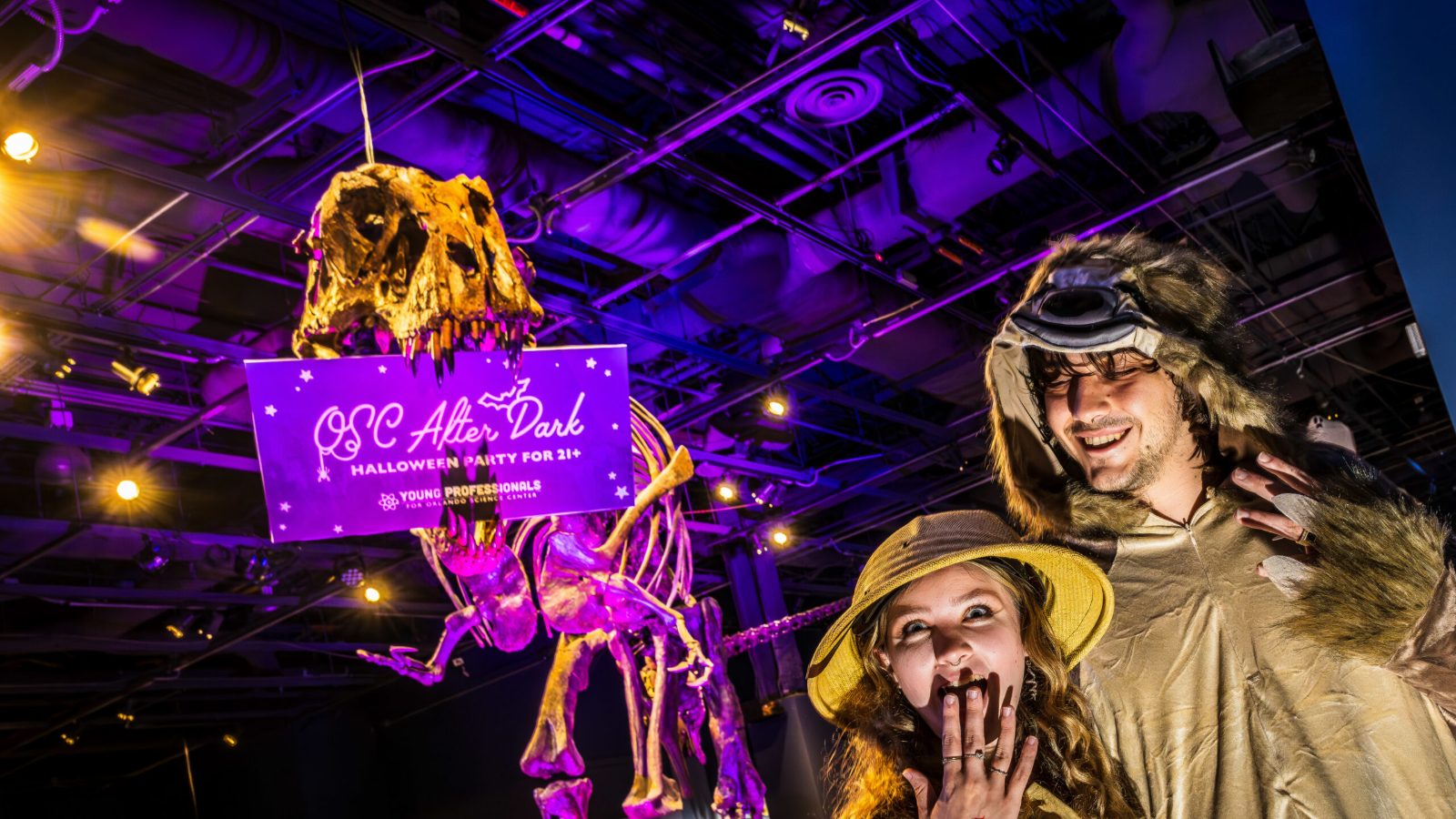 OSC After Dark 2026 guests in explorer costumes posing beneath a glowing dinosaur skeleton in DinoDigs at Orlando Science Center